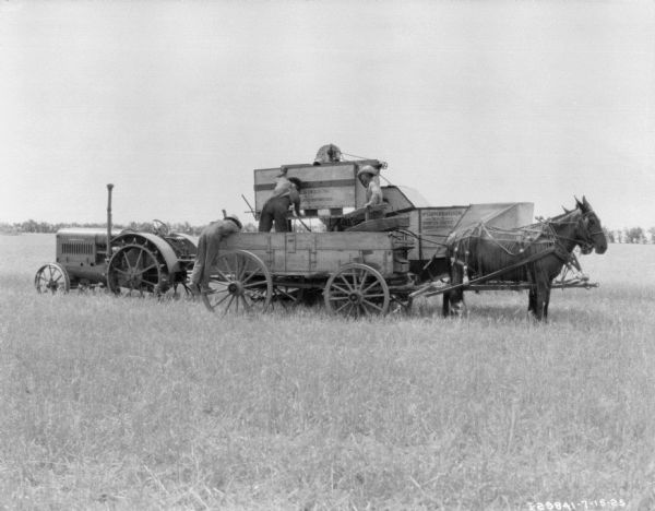 Left side view of a McCormick-Deering tractor attached to a harvester thresher parked in a field. Three men are working in a horse-drawn wagon parked in front of the harvester thresher. The horses are wearing fly-nets.