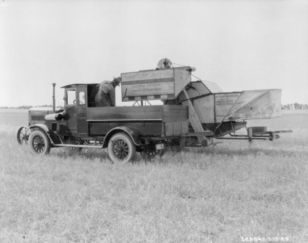 A man is standing in the bed of a truck in the foreground. Behind the truck is a McCormick-Deering tractor, which is pulling a McCormick-Deering No. 8 power drive harvester thresher.