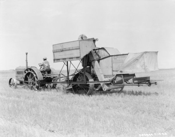 Three-quarter view from left rear of a man driving a tractor to pull a McCormick-Deering No. 8 power drive harvester thresher in a field.