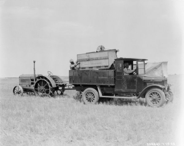 A man is sitting in the driver's seat of a truck parked in front of a tractor-drawn harvester thresher in a field. Another man is standing and working on the McCormick-Deering No. 8 power drive harvester thresher.