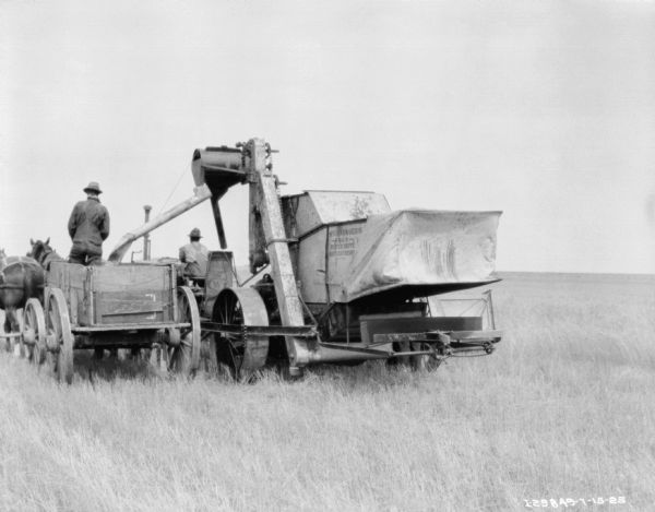 Rear view of a man on a McCormick-Deering tractor pulling a McCormick-Deering No. 8 power drive harvester thresher in a field. Alongside on the left is a man on a horse-drawn wagon drawn by two horses wearing fly-nets.