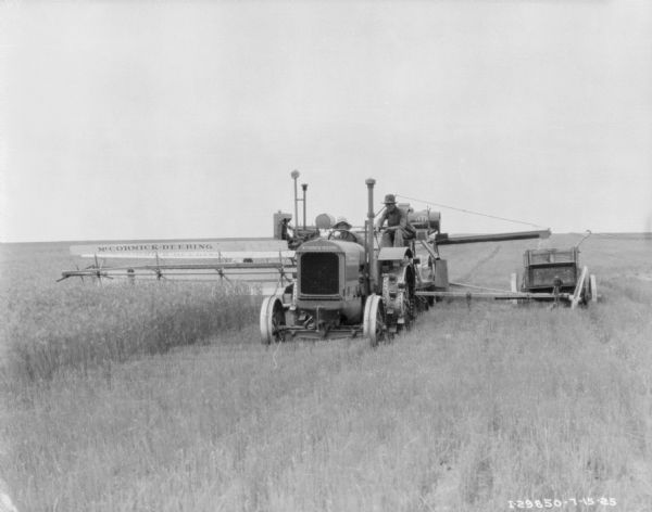 View from front of two men on a McCormick-Deering No. 8 power drive harvester thresher in a field. There is a wagon nearby on the right.