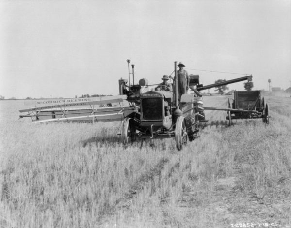 View from front of a man driving an International tractor pulling a McCormick-Deering harvester thresher. A man is standing on the harvester thresher, and a wagon is parked alongside on the right. Farm buildings and a water tower are in the far distance. 