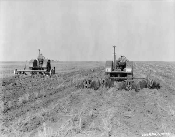 Rear view of a man on the left, and a young boy on the right, both driving McCormick-Deering 15-30 tractors with cultivators in a field.