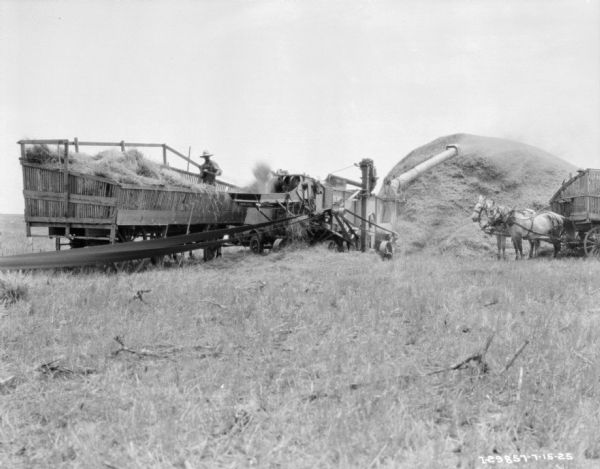 View across field towards a man standing and working in a wagon full of hay next to a thresher powered by a long belt, seen in the foreground. There is a pile of hay in the background on the right. There is a horse-drawn wagon on the far right.