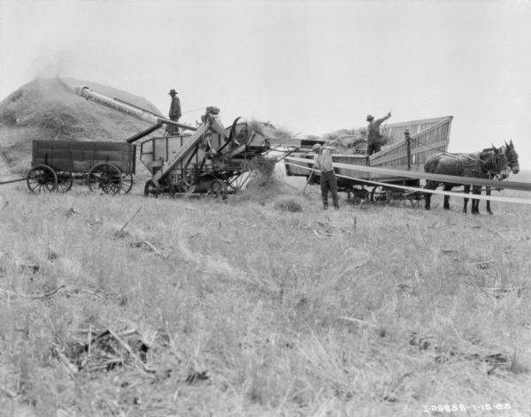View across field towards three men working with a McCormick-Deering threshing machine. One man is standing on the thresher near the chute, which is shooting grain onto a large pile of hay on the left. Another man is standing near the back of the thresher raking hay. The third man is standing in a horse-drawn wagon raking hay into the back of the thresher. A dog is standing under the wagon. The thresher is being powered by a belt which stretches from the right foreground to the threshing machine.
