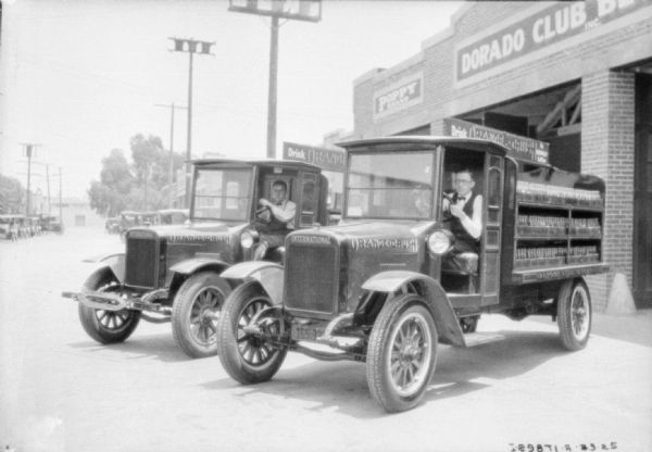 Two men are sitting in the driver's seats of two delivery trucks for Orange Crush. The beds of the trucks are open, and the truck on the right is carrying three shelves of wooden crates holding glass bottles. They are parked at an angle in front of a brick building with large open garage doors. Signs on the building read: "Dorado Club" and "Drink Orange Crush."