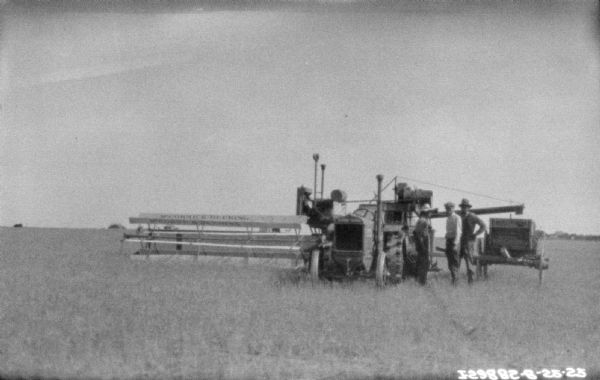 Three men are standing in a field near a tractor pulling a McCormick-Deering harvester thresher. There is a wagon on the right.