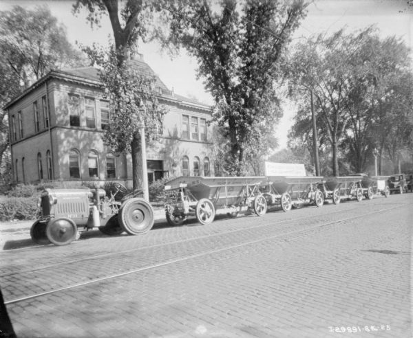 View across street towards a McCormick-Deering No. 11 tractor and four wagons parked along the opposite curb. A sign on one of the wagons reads: "McCormick-Deering Industrial Tractors, Help Keep Maywood Clean."