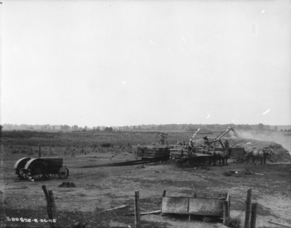 Elevated view of a threshing operation. A tractor on the left is powering the thresher with a long belt. Three men are standing in three horse-drawn wagons while pitching hay into the thresher.