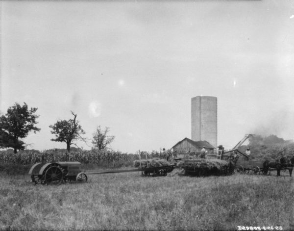 View across field towards threshing operation. A tractor on the left is powering the thresher with a long belt. Three men are standing in three horse-drawn wagons while pitching hay into the thresher. In the background is a cornfield, farm buildings, and a silo.