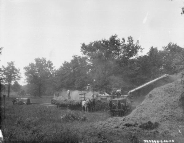 Elevated view across field towards a threshing operation. There is a tractor on the left powering the thresher with a long belt. Three men are standing in three horse-drawn wagons while pitching hay into the thresher. A large pile of hay is on the right.