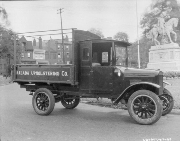 Right side view of the Kalada Upholstering Co. delivery truck parked on a street. Buildings are in the background on the left. Behind the truck on the right is a monument with a man riding a horse,