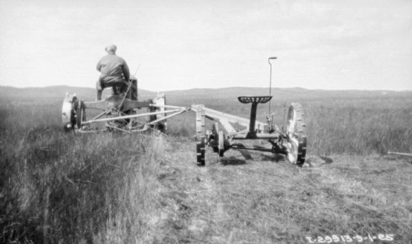 Rear view of a man on a Farmall tractor pulling a mower in a field.