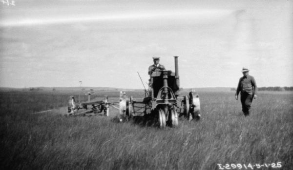View from front of a man driving a farmall to pull a mower in a field. A man is standing nearby on the right.