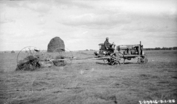 Profile view of a man on a Farmall tractor pulling a dump rake in a field. There is a tall, conical stack of hay in the distance.