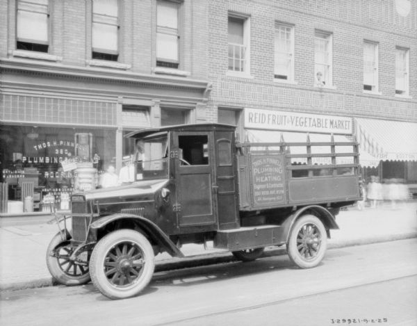 View across street towards a delivery truck parked at a curb. Behind the truck are storefronts along a sidewalk. A sign on the side of the truck reads: "Thos. H. Pinnell, Plumbing and Heating."