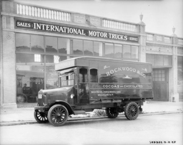 View across street towards a truck parked along the curb in front of an International dealership. A sign on the truck reads: "Rockwood & Co., Cocoas and Chocolates."