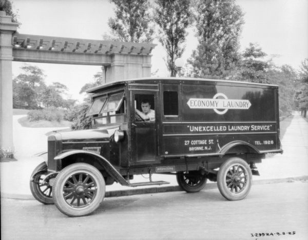 View across road towards a man sitting in the driver's seat of a delivery truck. A sign on the side of the truck reads: "Economy Laundry." Behind the truck are columns and an archway into what may be a park.