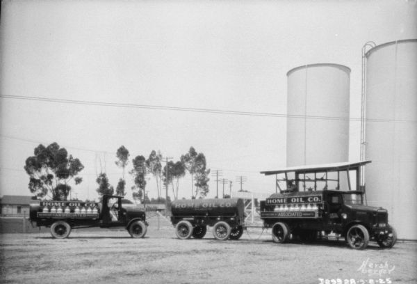 Two oil delivery trucks and a tank on a trailer for Home Oil Co. parked in front of oil storage tanks. 