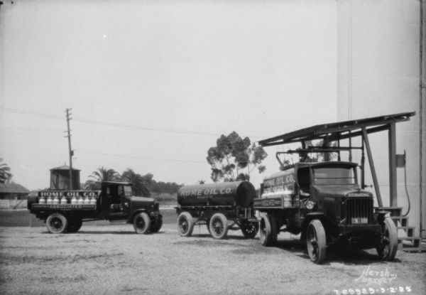 Two oil delivery trucks and a tank on a trailer for Home Oil Co. parked in front of oil storage tanks. 