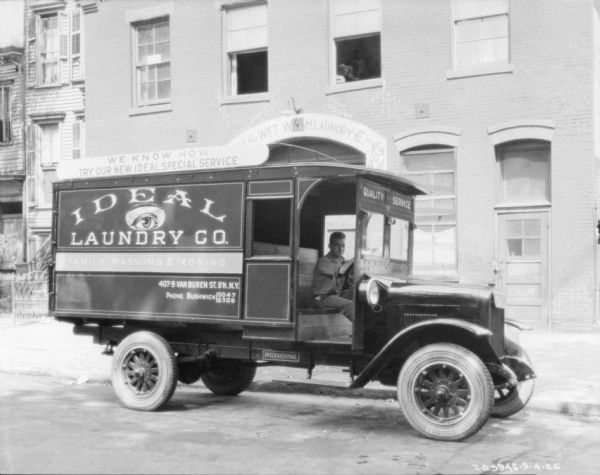 View across street towards a man sitting in the driver's seat of an Ideal Laundry Co. truck parked along a curb. Behind the truck is a building with an arched entrance, and the sign in the arch reads, in part: "Wet W.H. Laundry Co."
