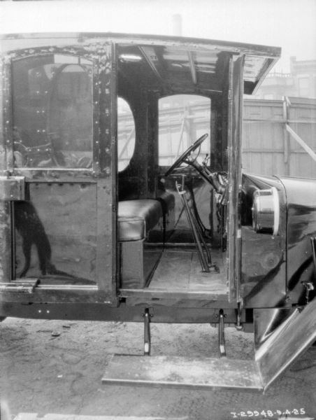 View through open passenger side door into cab of truck showing foot pedals. Behind the truck is a tall, wooden fence, with brick buildings in the background.