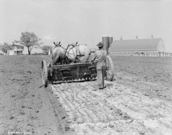 Rear view of a man walking behind a McCormick-Deering lime sower in a field. In the distance is a farmhouse on the left, and barns on the right.