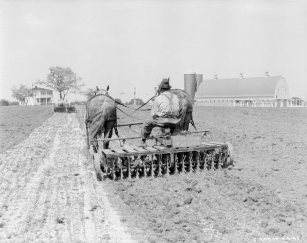 Rear view of a man riding a horse-drawn rotary hoe in a field. Another man in the distance is walking behind a horse-drawn lime sower.