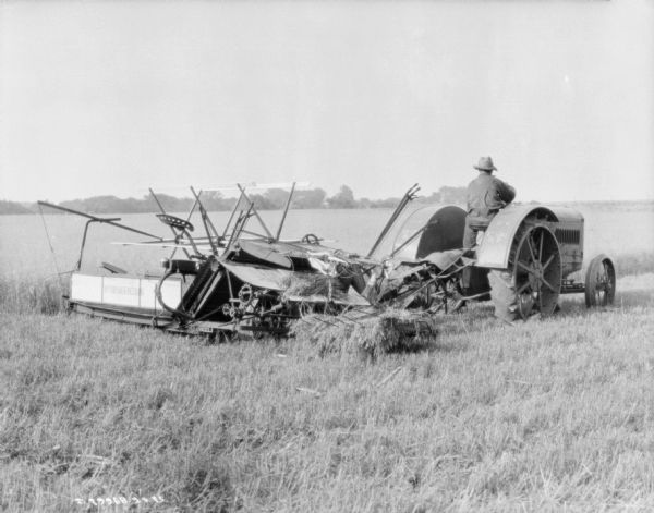 Three-quarter view from right rear of a man driving a 15-30 tractor pulling a McCormick-Deering binder in a field.