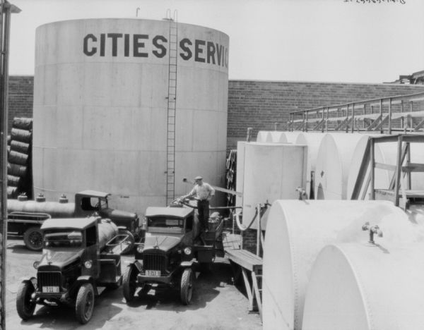 Elevated view of three delivery trucks parked at oil tanks. A sign on the largest tank reads, in part: "Cities Servic_." A man is filling up one of the tanks on the trucks from one of the storage tanks.
