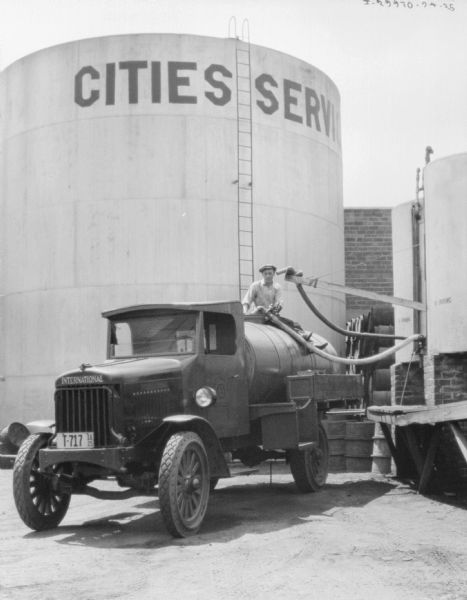 A man is filling an oil delivery truck from a storage tank. A sign on the largest tank reads, in part: "Cities Ser..."