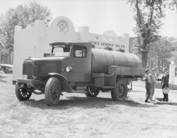 Three-quarter view from front left of a Cities Service Oil Company delivery truck parked outdoors. In the background is a building with a sign for "Cities Service Oils." Three men are standing at the back of the truck.