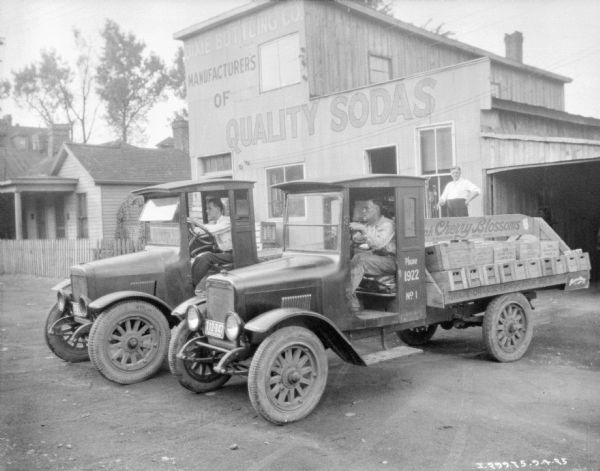 Slightly elevated view of two men sitting in the driver's seats of two delivery trucks parked in front of a building with a sign that reads: "Dixie Bottling Co., Manufacturer's of Quality Sodas. A man is standing near the doorway of the building."