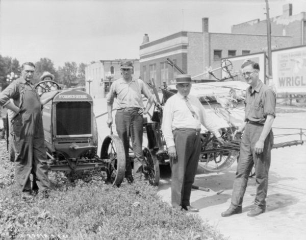 Group of men posing outdoors with a McCormick-Deering binder sitting on a street. There is a child sitting in the tractor seat of a McCormick-Deering sitting in the grass next to the binder. Across the street are commercial buildings and billboards.