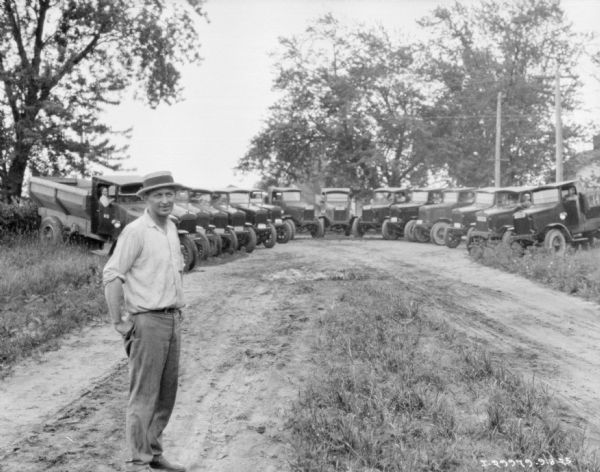 A man is standing on a dirt road in the foreground. In the background is a fleet of trucks, approximately 15, parked in a semicircle at the end of the road near trees.