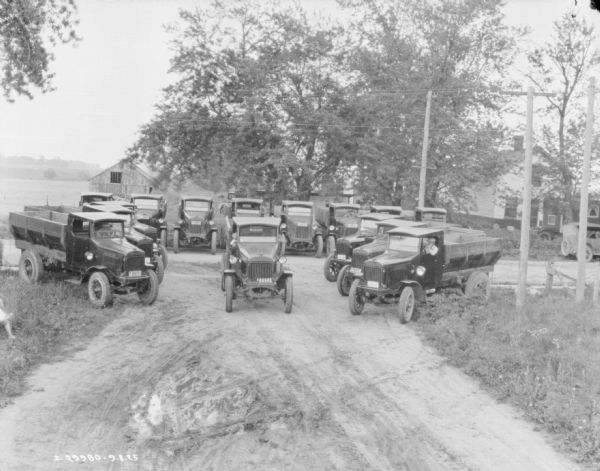 Slightly elevated view of a fleet of trucks, approximately fifteen, parked in a square formation at the end of a dirt road. There appear to be men sitting in the driver's seat of every truck. Buildings are in the background.