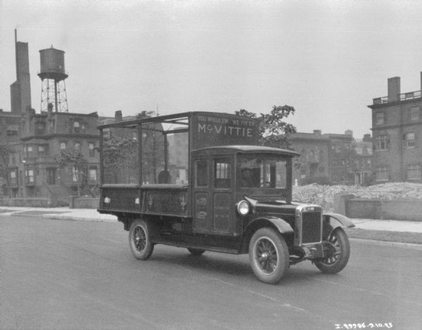 View from side of road towards a man in the driver's seat of a delivery truck on a street. The sign on the front of the truck above the cab reads: "You Wreck Em, We Fix Em." There are buildings and a water tower in the background.