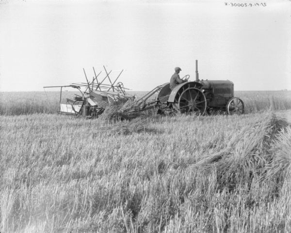 View across field towards a man driving a tractor to pull a binder. Bundles of wheat are lying in the field.