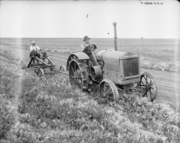 Three-quarter view from front right of a man driving a McCormick-Deering tractor, pulling a man on a plow in a field.