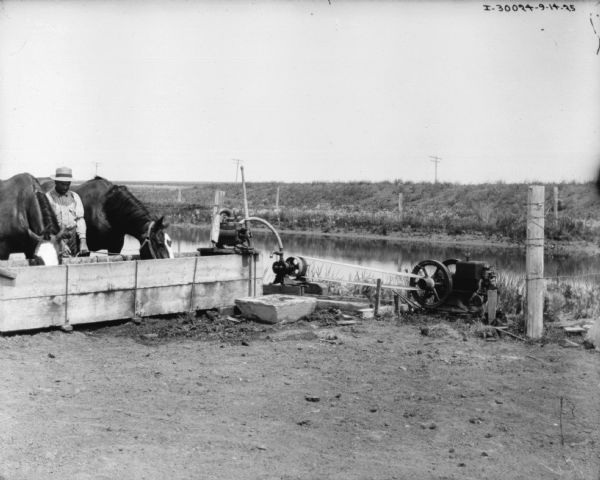 A man is standing between two horses who are drinking water from a trough filled by a pump. There is a body of water in the background. 