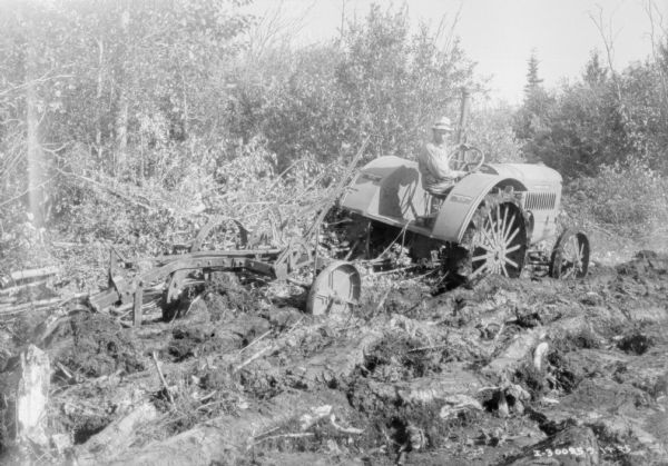 Rear view of a man plowing rough land with a McCormick-Deering 15-30 tractor near forest.
