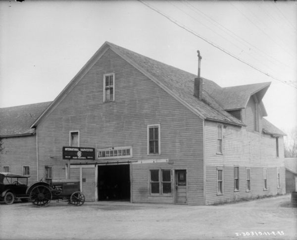 View of building with an International Harvester sign above an open garage door. An automobile and a tractor are parked on the left.