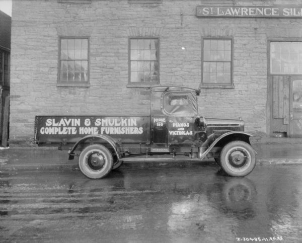 Side view of a truck parked alongside a stone building. A sign on the building reads in part: "St. Lawrence ..." The signs painted on the truck read: "Slavin & Shulkin Compete Home Furnishers: and "Pianos and Victrolas."