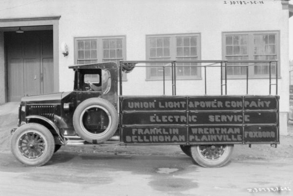 Left side view of man in the driver's seat of a truck parked in front of a building. The sign painted on the side of the truck bed reads: "Union Light & Power Company, Electric Service, Frankin Wrentham, Bellingham, Plainville, Foxboro."