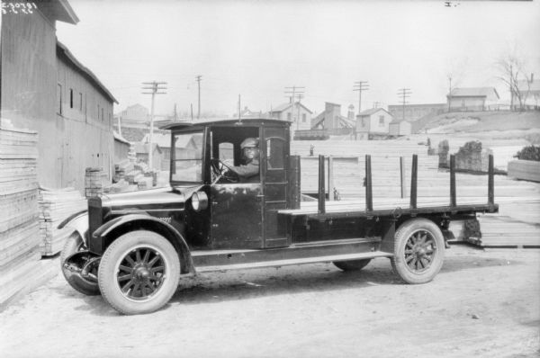 View of a man sitting in the driver's seat of a truck parked near a building at a lumberyard. There are houses in a neighborhood in the background.