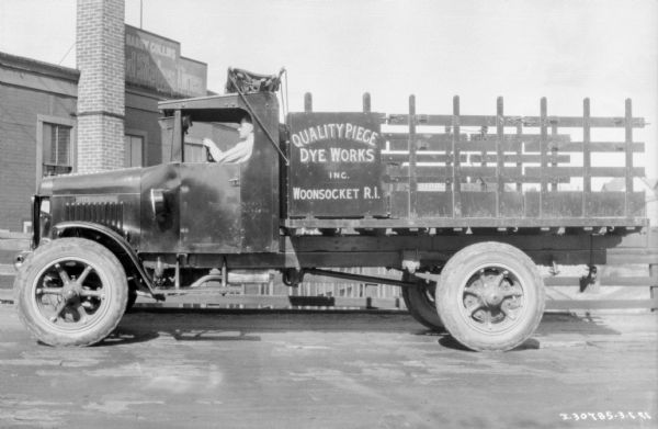 Left side view of a man sitting in the driver's seat of a delivery truck near a building with a sign that reads: "Harry Collins United States Tires." The sign on the truck reads: "Quality Piece Dye Works Inc. Woonsocket R.I."