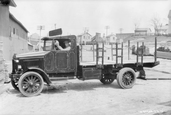 Left side view of a man sitting in the driver's seat of a delivery truck near a building with lumber stacked in front of it. There are houses in a neighborhood in the background.