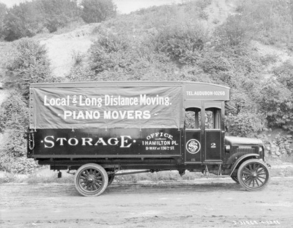 International truck parked on a dirt road. Signs on the side of the truck read: "Local & Long Distance Moving," "Piano Movers," and "Storage." In the background is a hill with trees and bushes.