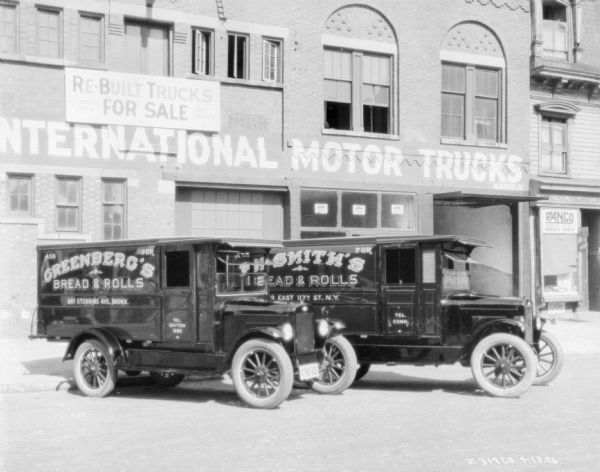 Two delivery trucks are parked at an angle in front of a two-story brick building. Signs on the building read: "Re-Built Trucks For Sale," and "...nternational Motor Trucks." The sign on the truck on the left reads: "Greenberg's Bread & Rolls." The sign on the truck on the right reads: "Smith's Bread & Rolls."